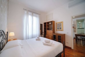 A hotel room with a bed, a nightstand with a lamp, a wooden cabinet, a dining area with chairs, and a painting on the wall.