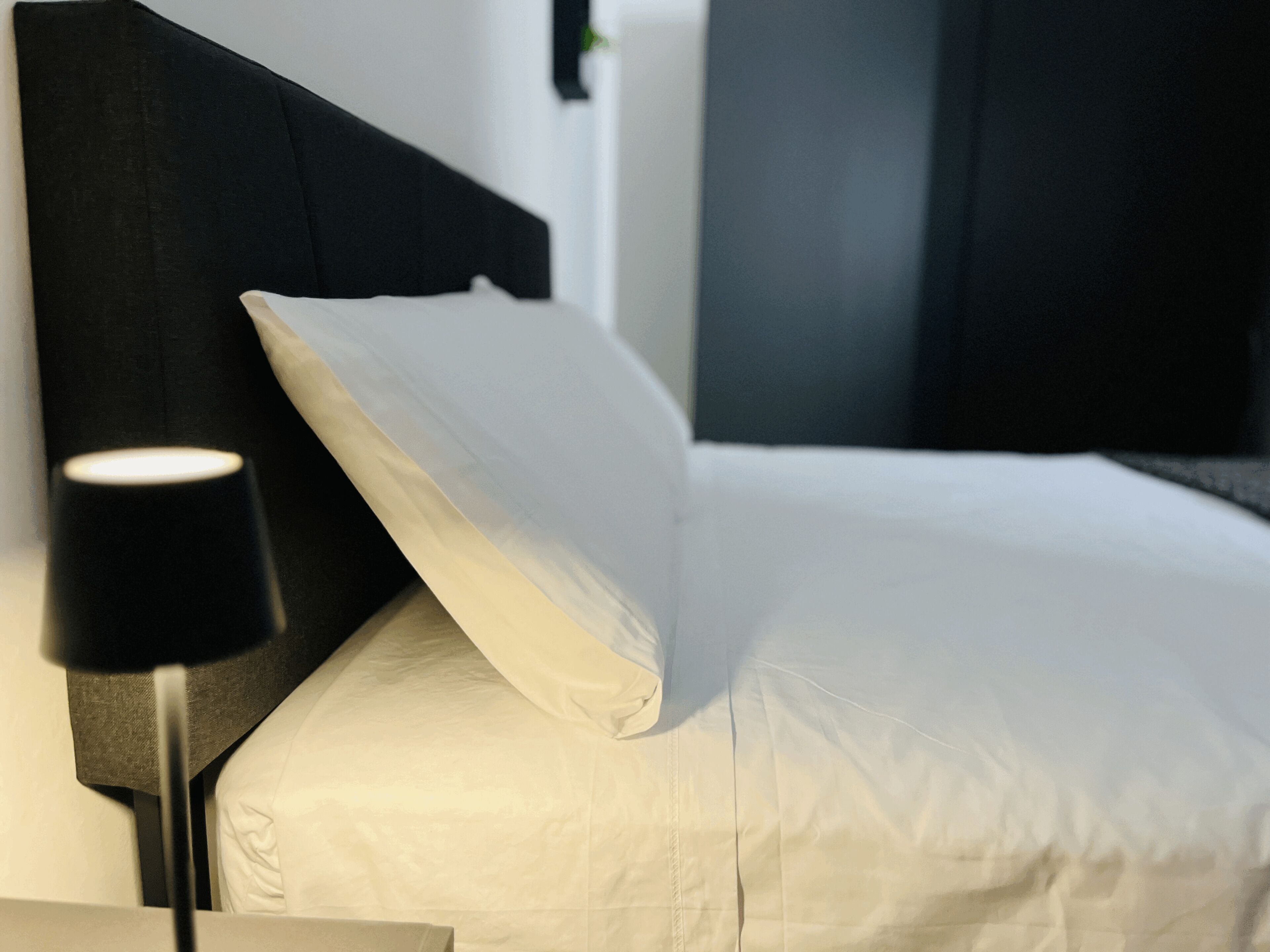 Een net opgemaakt bed met witte lakens, een zwart hoofdbord en een bedlampje.