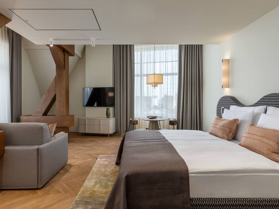 A modern hotel room with a large bed, a sofa, a TV, and a dining area with a table and chairs.
