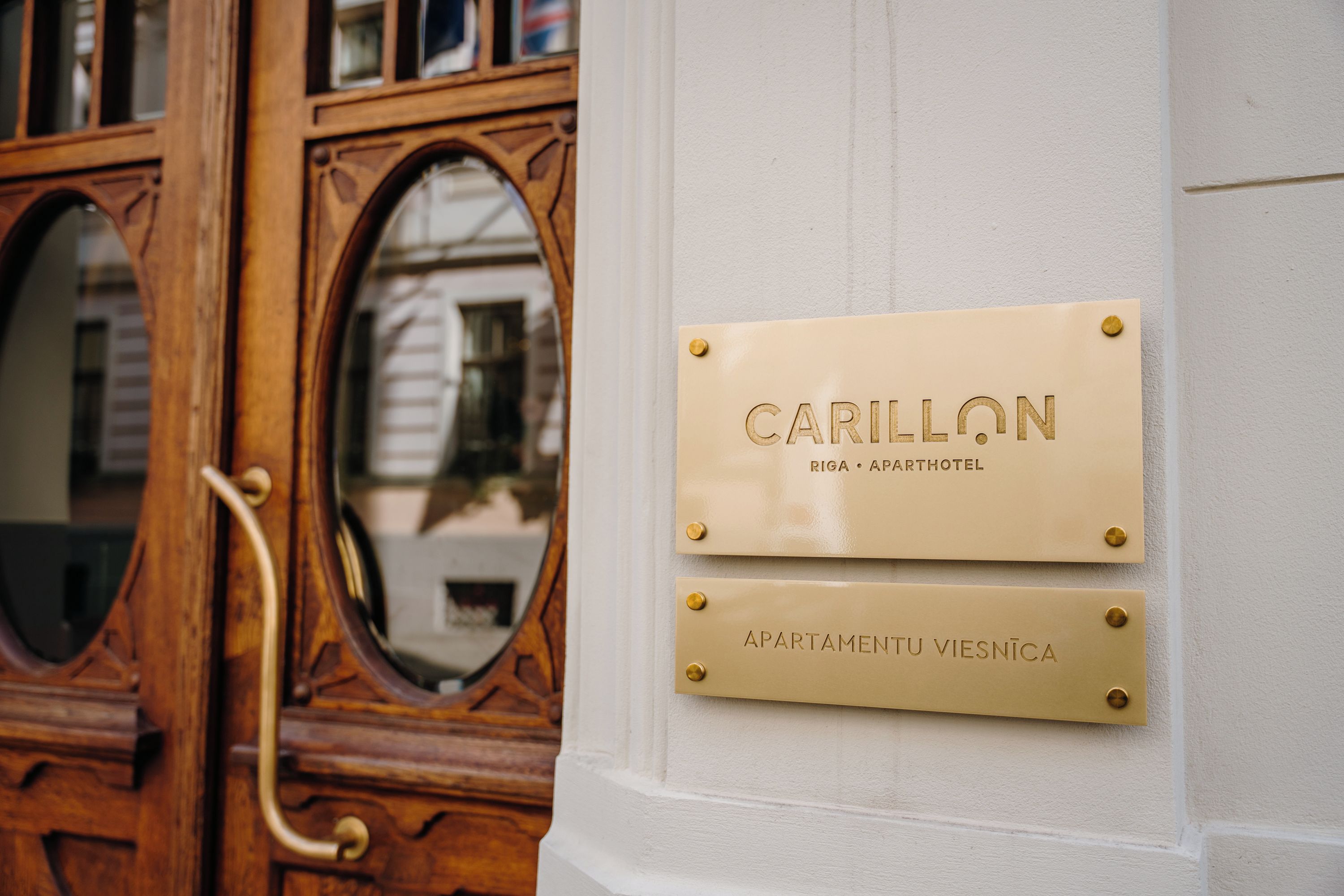 Una puerta de madera con picaporte de latón y un panel de vidrio. Hay dos placas en una pared, una dice 'CARILLON RIGA APARTHOTEL' y la otra 'APARTMENTU VIESNICA'.