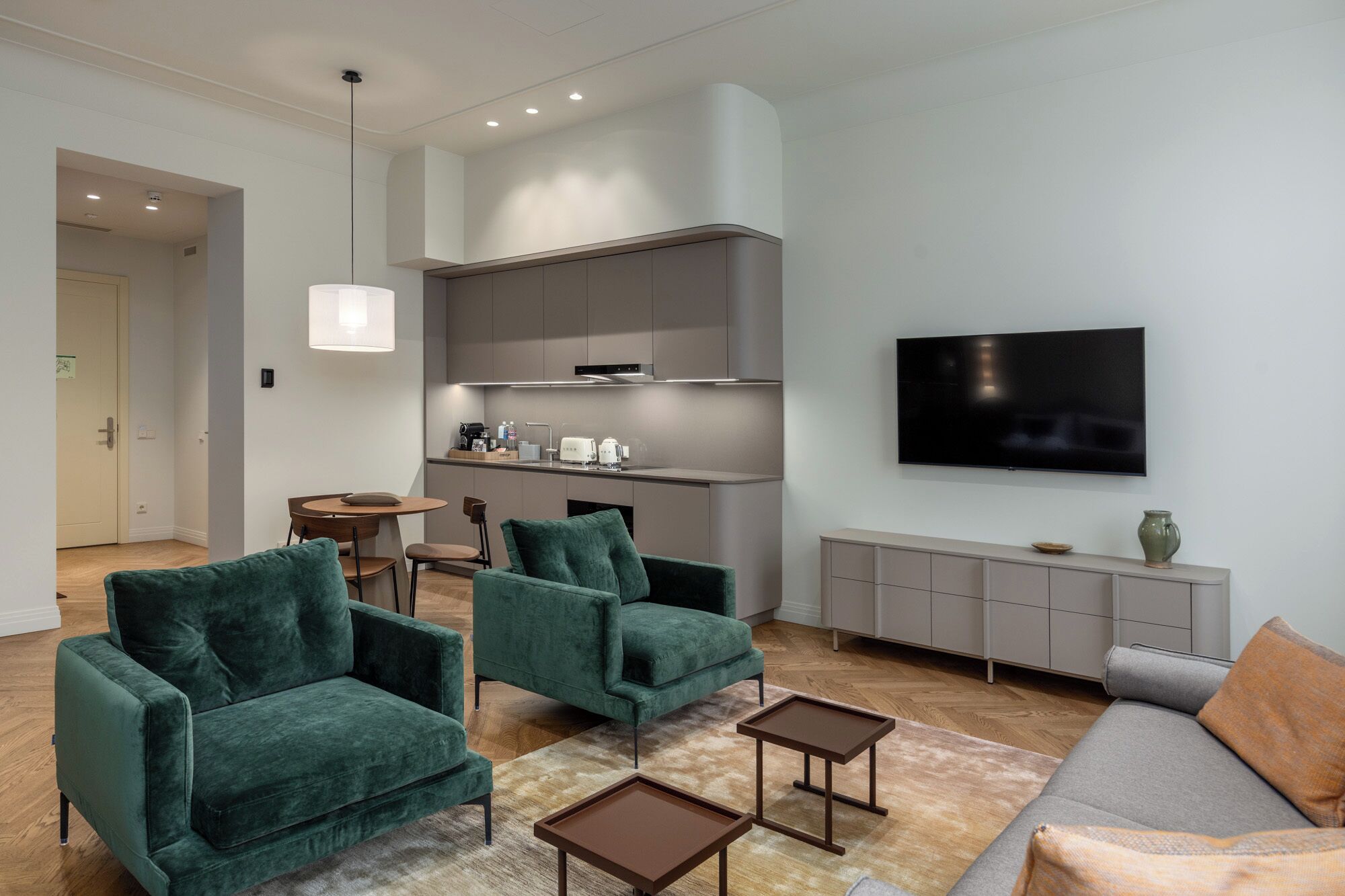 A modern living room with two green armchairs, a grey sofa, a coffee table, a TV, and a kitchen area with a built-in oven and microwave.