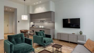 A modern living room with two green armchairs, a grey sofa, a coffee table, a TV, and a kitchen area with a built-in oven and microwave.