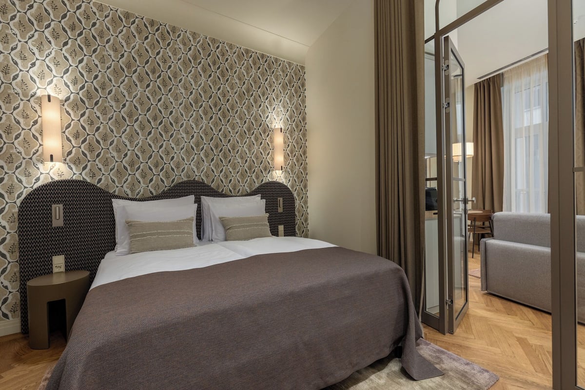 Une chambre d'hôtel moderne avec un papier peint à motifs, un grand lit doté d'une tête de lit sombre, des tables de chevet et une vue sur un balcon, accessible par une porte vitrée.