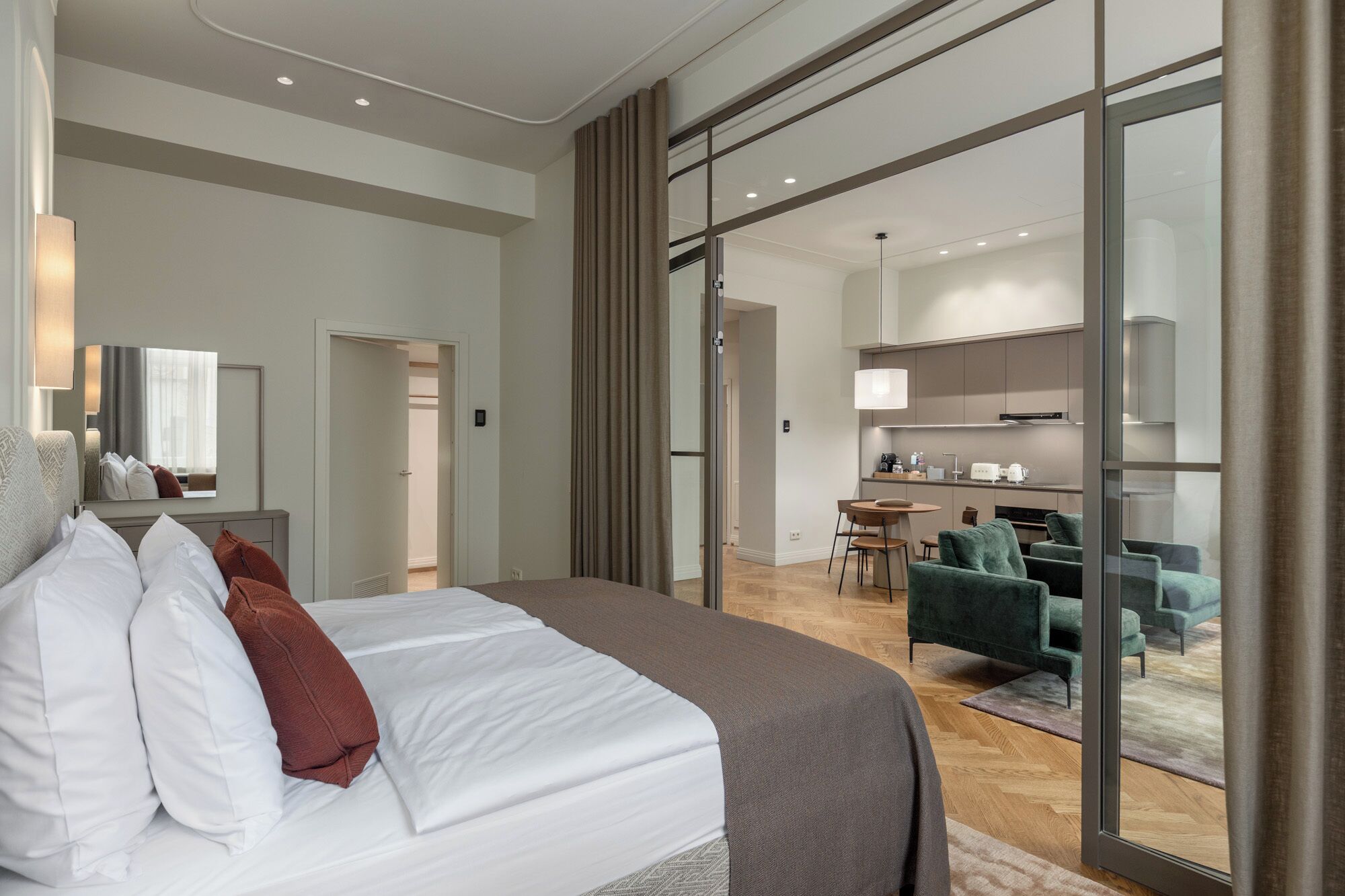A modern hotel room with a large bed, a separate dining area, and a kitchen visible through a glass door.