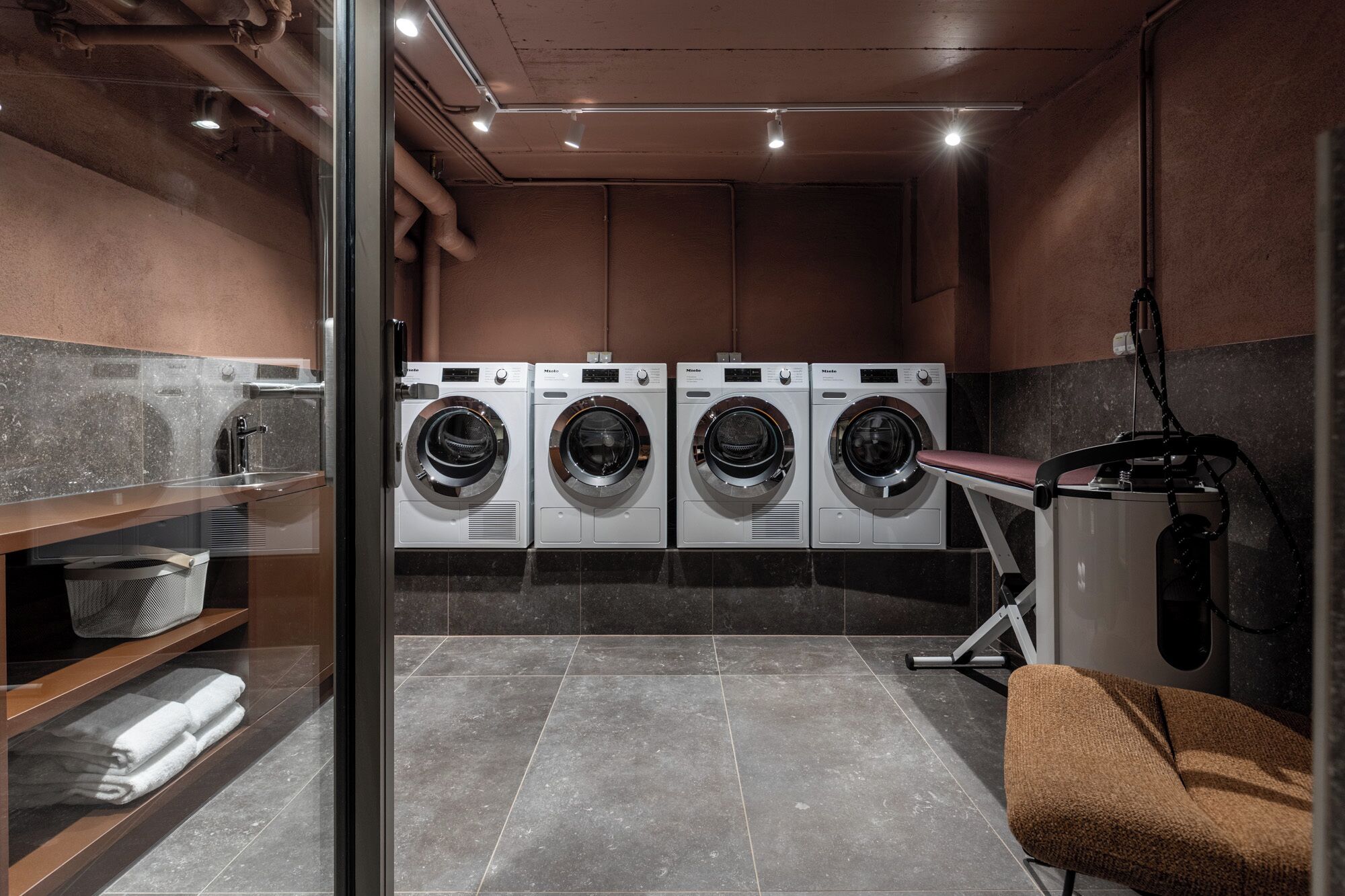 A laundry room with four front-loading washers and dryers, a towel rack, and a bench.