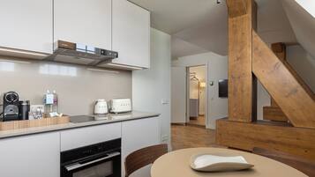 A modern kitchen with white cabinets, a built-in oven, and a coffee machine. There's a wooden staircase and a dining area with a table and chairs.