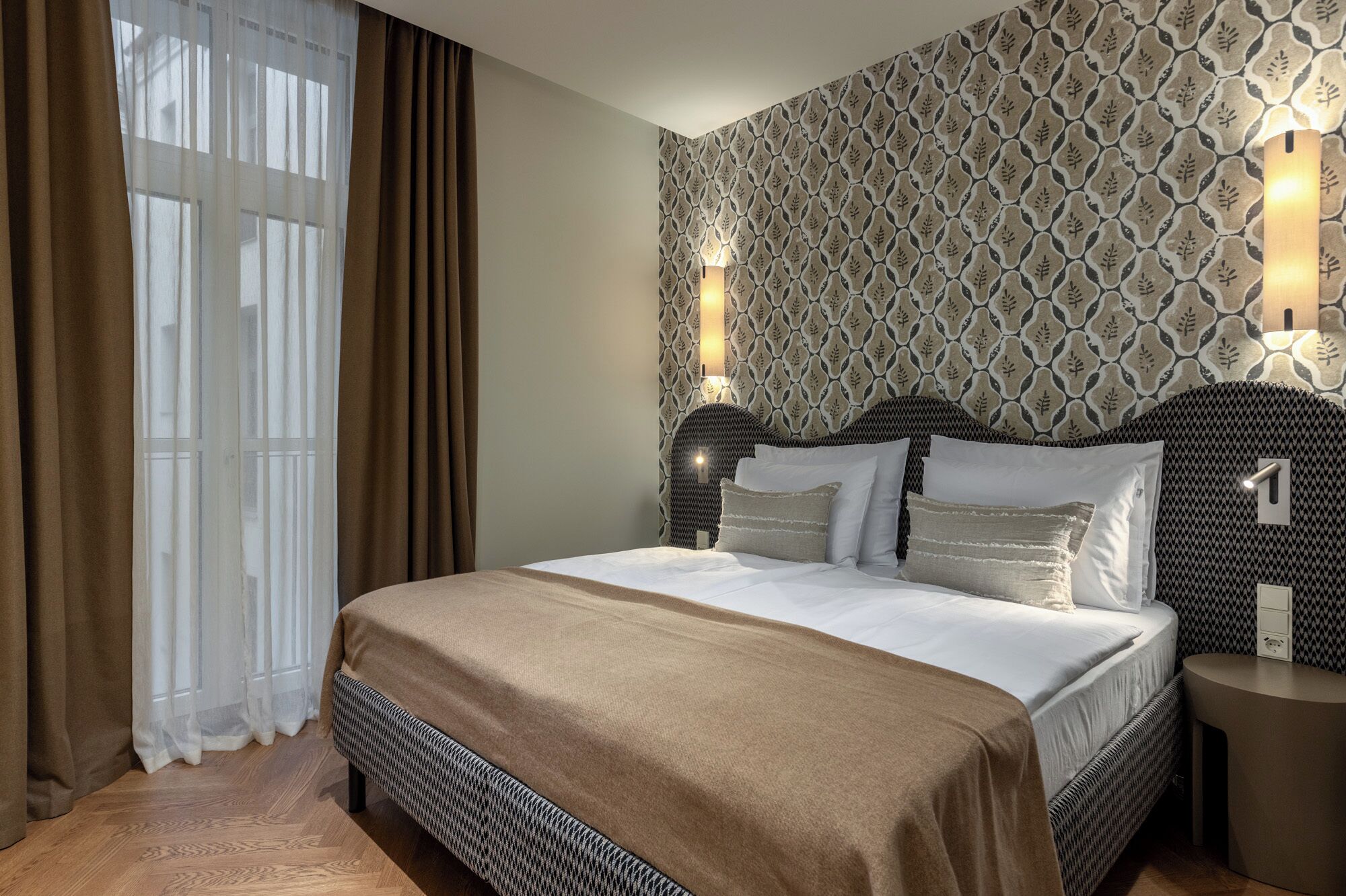 A hotel room with a large bed, two bedside tables, a patterned wallpaper, and a window with curtains.