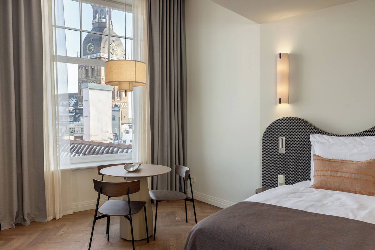 Une chambre d’hôtel avec un lit, une table ronde, deux chaises, une fenêtre avec des rideaux et une vue sur la ville.