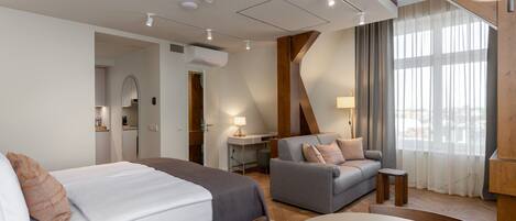 A modern hotel room with a bed, a sofa, a table, and a view of the city through large windows.