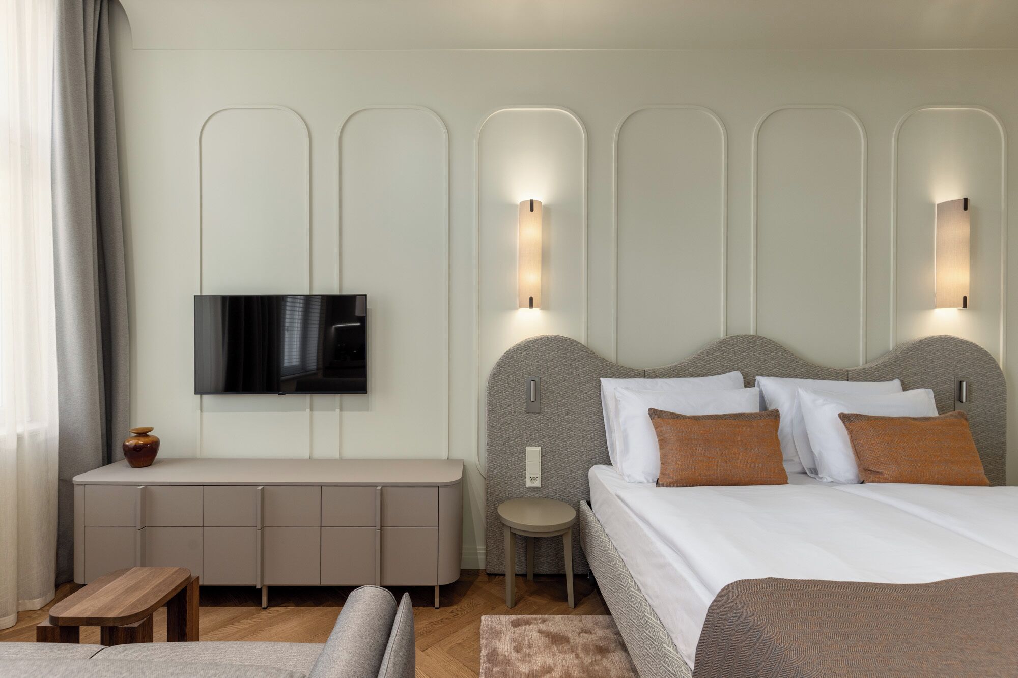 Ein modernes Hotelzimmer mit an der Wand montiertem Fernseher, einem Bett mit vielen Kissen, einem Nachttisch und einem kleinen Hocker.