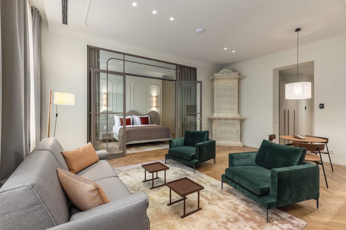 Une chambre d’hôtel moderne avec un canapé gris, des fauteuils verts, une petite table, un lit séparé par une cloison en verre et une cheminée.