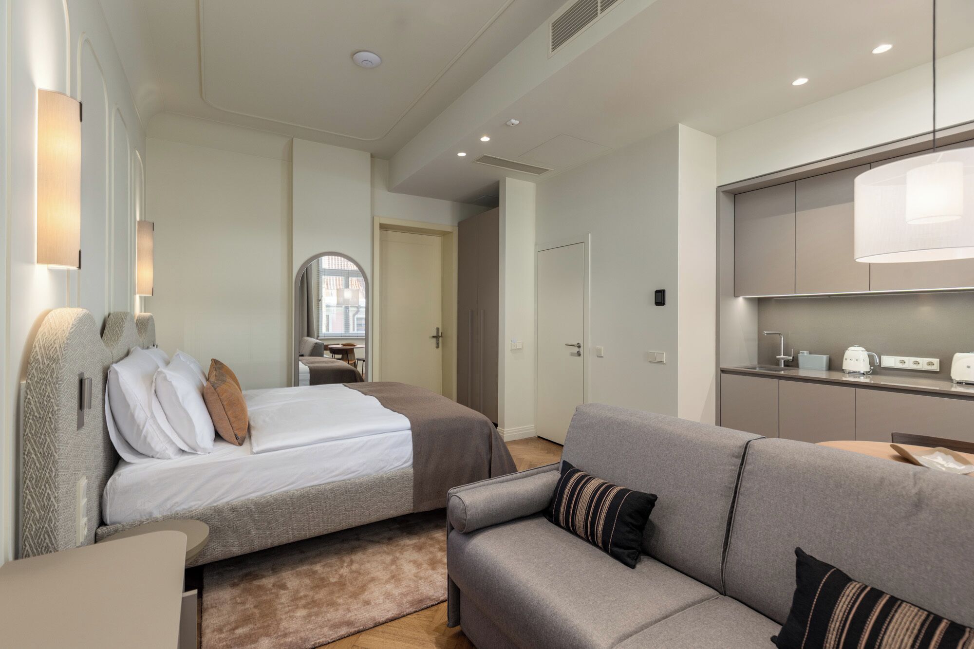 A modern hotel room with a bed, a sofa, a small table, and a kitchenette.