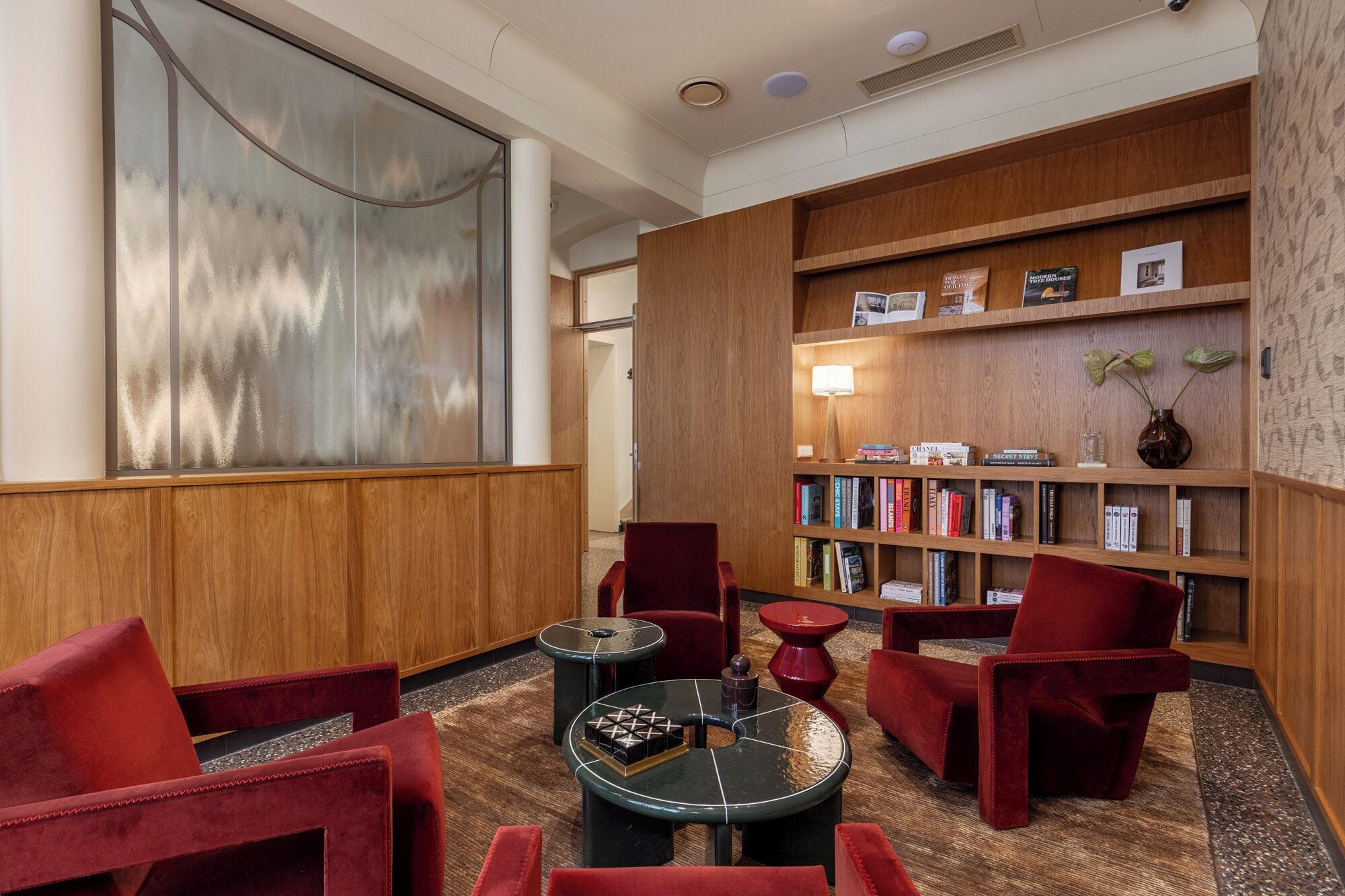 Un lobby moderno con una estantería de madera, sillas tapizadas en rojo, una mesa redonda y un cuadro abstracto grande.
