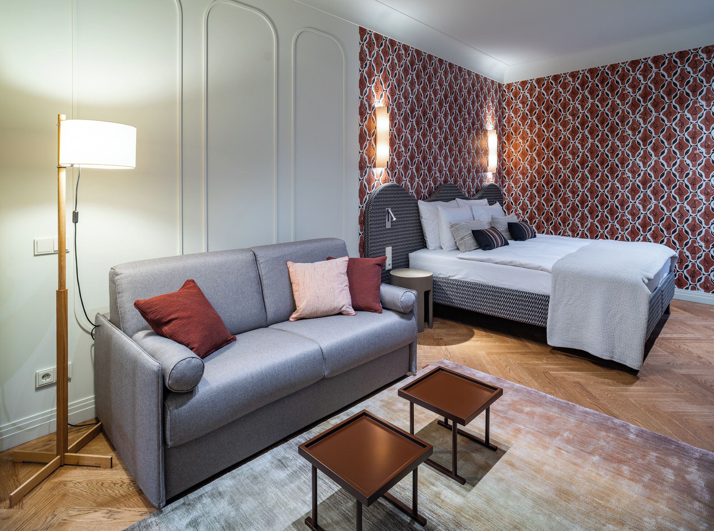 Una habitación de hotel moderna con un sofá gris, una cama con ropa de cama estampada, una mesita redonda pequeña y una lámpara de pie.
