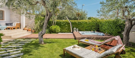 A backyard with a hot tub, lounge chairs, a wooden deck, and a stone wall.