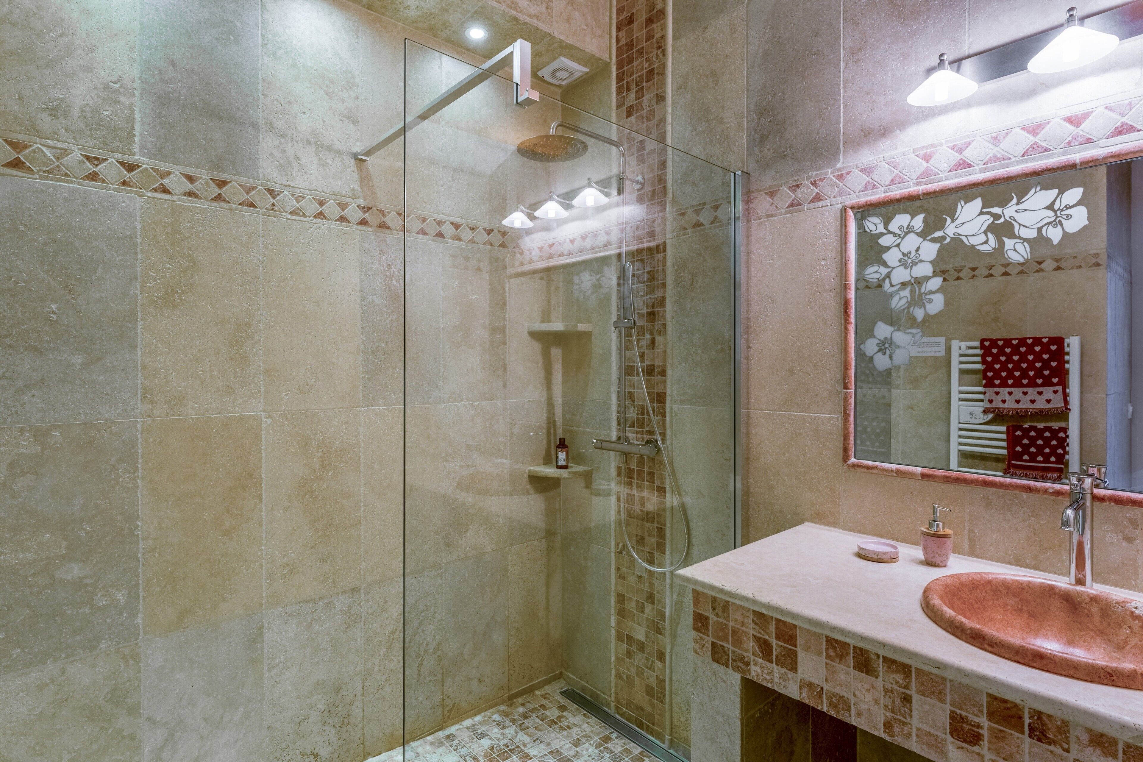Un baño con ducha envuelta en vidrio, un lavamanos de cobre y un espejo con diseño floral.