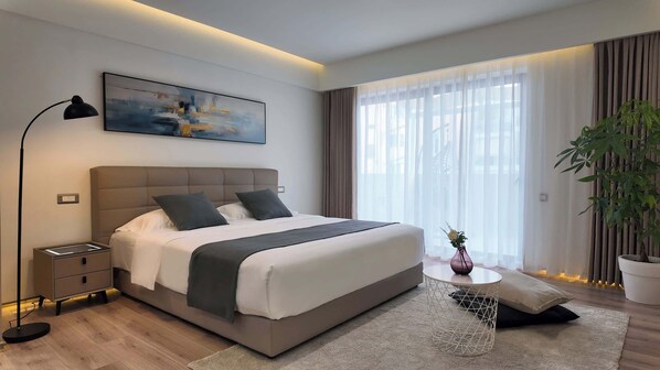 A modern hotel room with a large bed, bedside tables, a floor lamp, a painting, a window with sheer curtains, and a potted plant.
