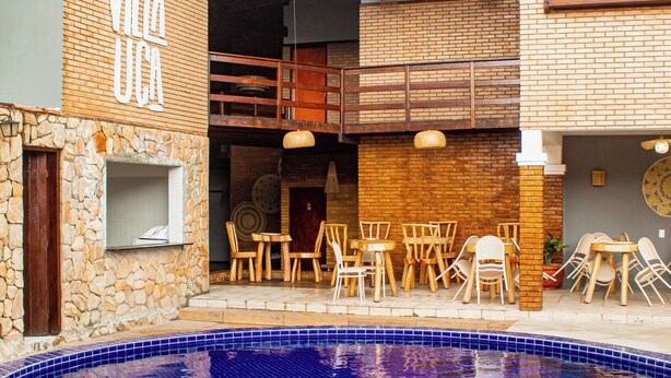 A pool area with a tiled edge, wooden furniture, and a building with a visible logo. - villa uca frances (Marechal Deodoro)