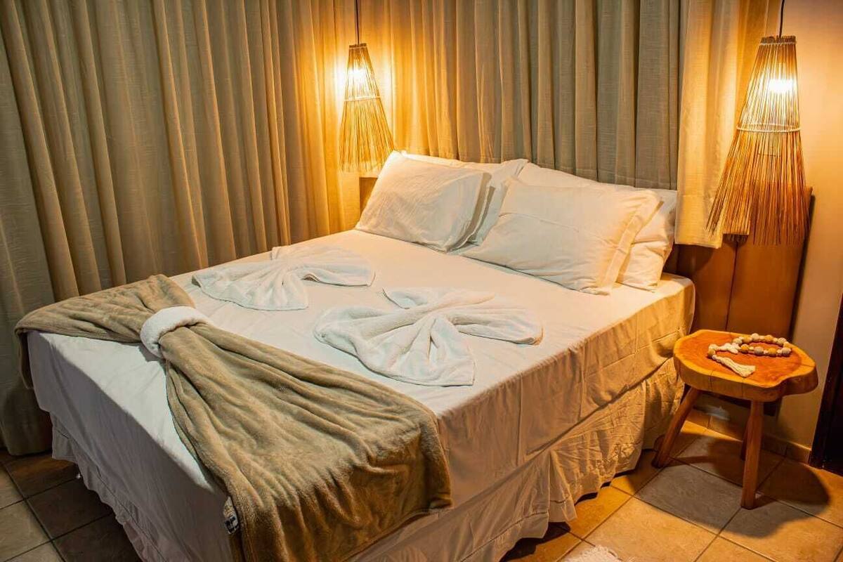 Cama bem arrumada com lençóis brancos e manta marrom, um banquinho de madeira com uma toalha, e duas luminárias fixadas na parede.