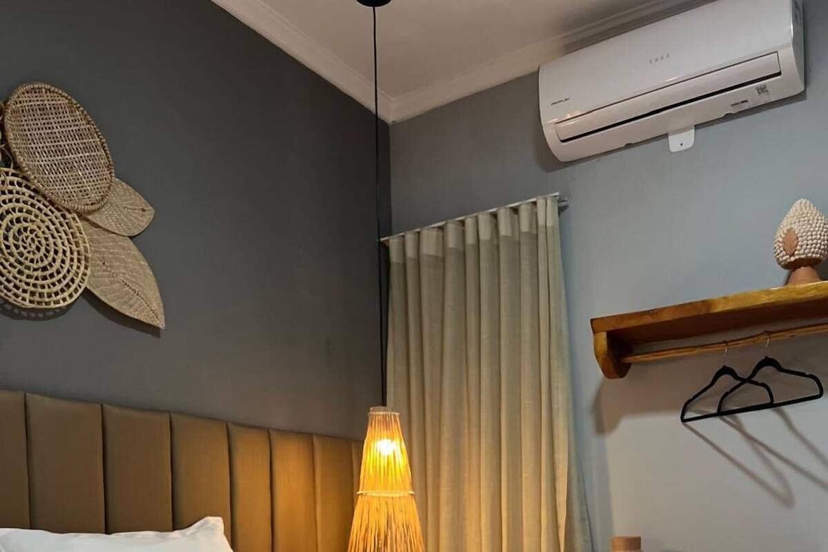 Quarto de hotel com cama, luminária pendente, janela com cortinas e ar condicionado.
