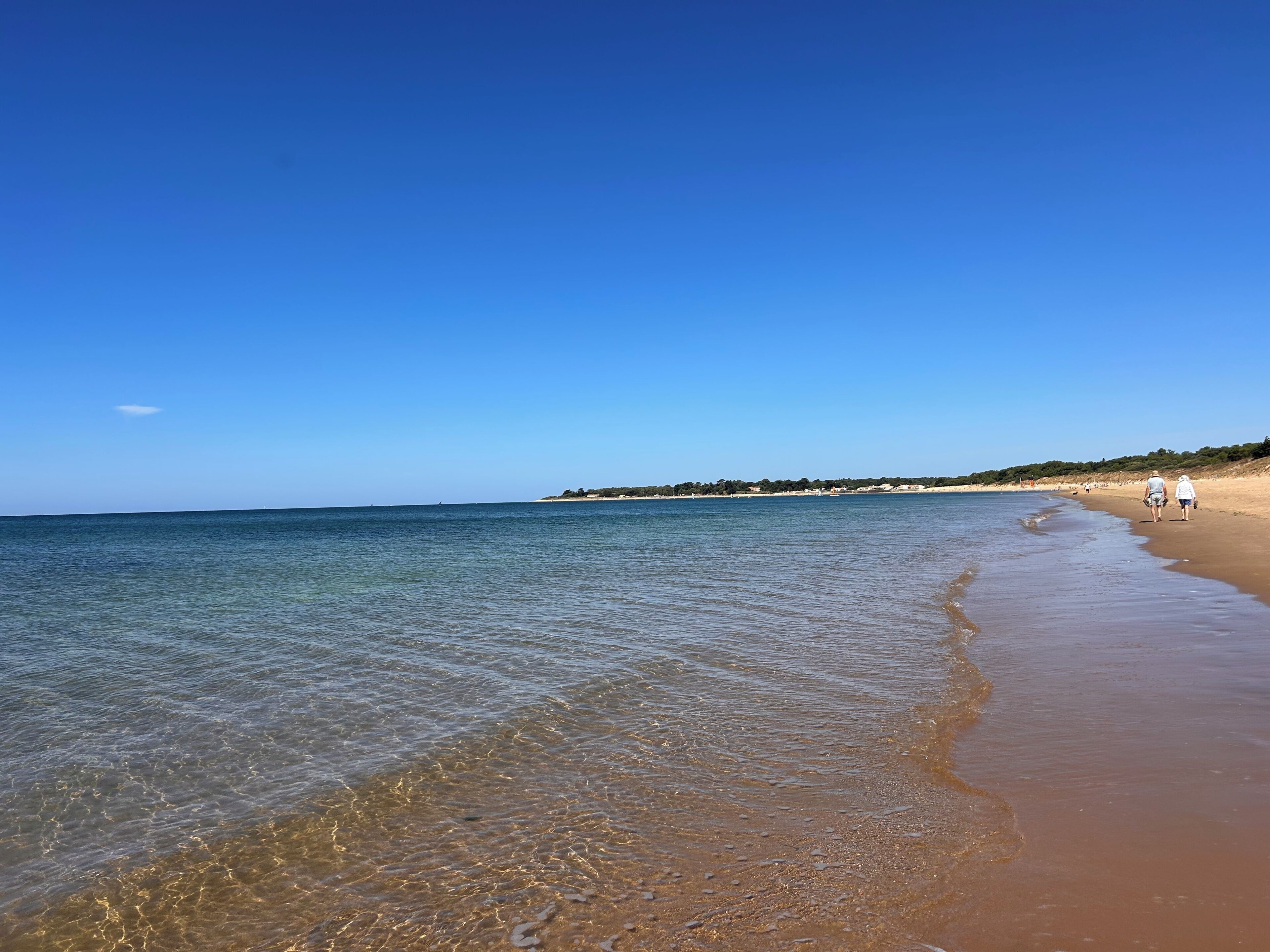 A beach with clear water, a sandy shore, and a few people walking.
