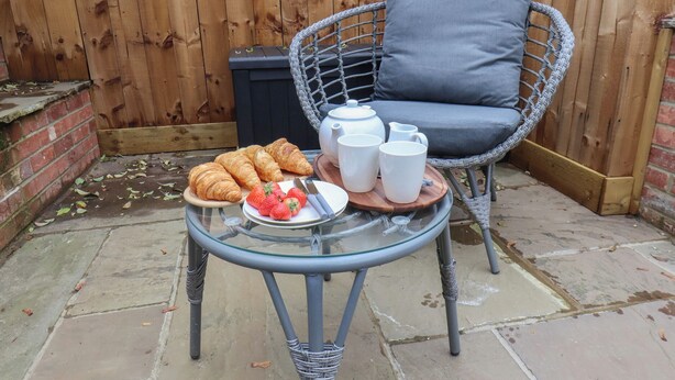 Une table en verre sur laquelle sont disposés des croissants, des fraises et des tasses blanches, sur une terrasse agrémentée d’une chaise en osier et d’un coussin gris.