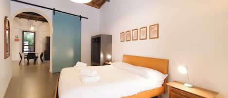 A hotel room with a bed, bedside table, and a desk with a chair. There is a hallway leading to another room.