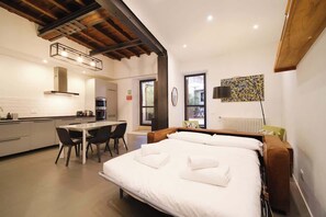 A modern hotel room with a large bed, a dining area, a kitchen, and a view of the outdoors.