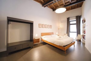 A bedroom with a bed, bedside tables, a wardrobe, a reading chair, a bookshelf, and a window with curtains.