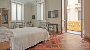 A spacious bedroom with a large bed, a desk, and a chair, a television mounted on the wall, and a balcony with a view of a yellow building. - SUYTE Torino Centro (Turin)