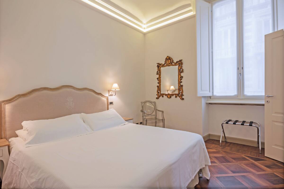 A neatly made bed with white linens, a wooden headboard, a wall-mounted mirror, a chair, and a small bench by a window with white curtains.