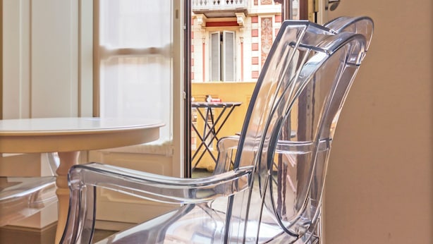 A clear glass chair and table in a room with wooden flooring, a white door, and a view of a building with balconies through an open door. - SUYTE Torino Centro (Turin)