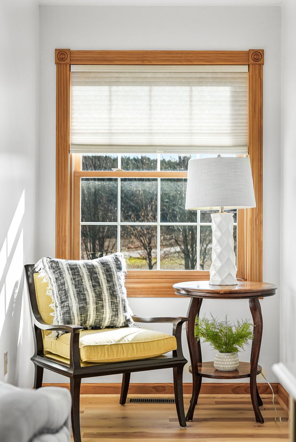 Una habitación con ventana, una silla con cojín amarillo, una mesa con lámpara y una planta en maceta.
