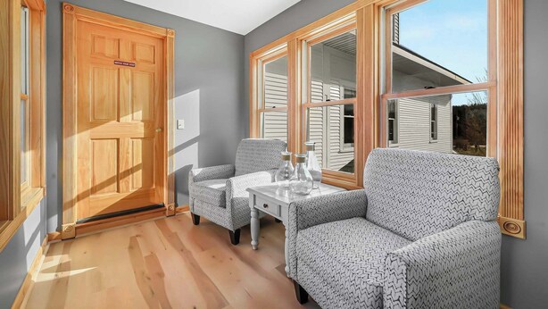 Une pièce dotée d’une porte en bois, de deux fauteuils à motifs, d’une petite table sur laquelle sont posées des bouteilles, et de grandes fenêtres offrant une vue sur une maison.