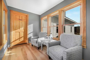 Ein Zimmer mit Holztür, zwei gemusterten Sesseln, einem kleinen Tisch mit Flaschen und großen Fenstern mit Blick auf ein Haus.