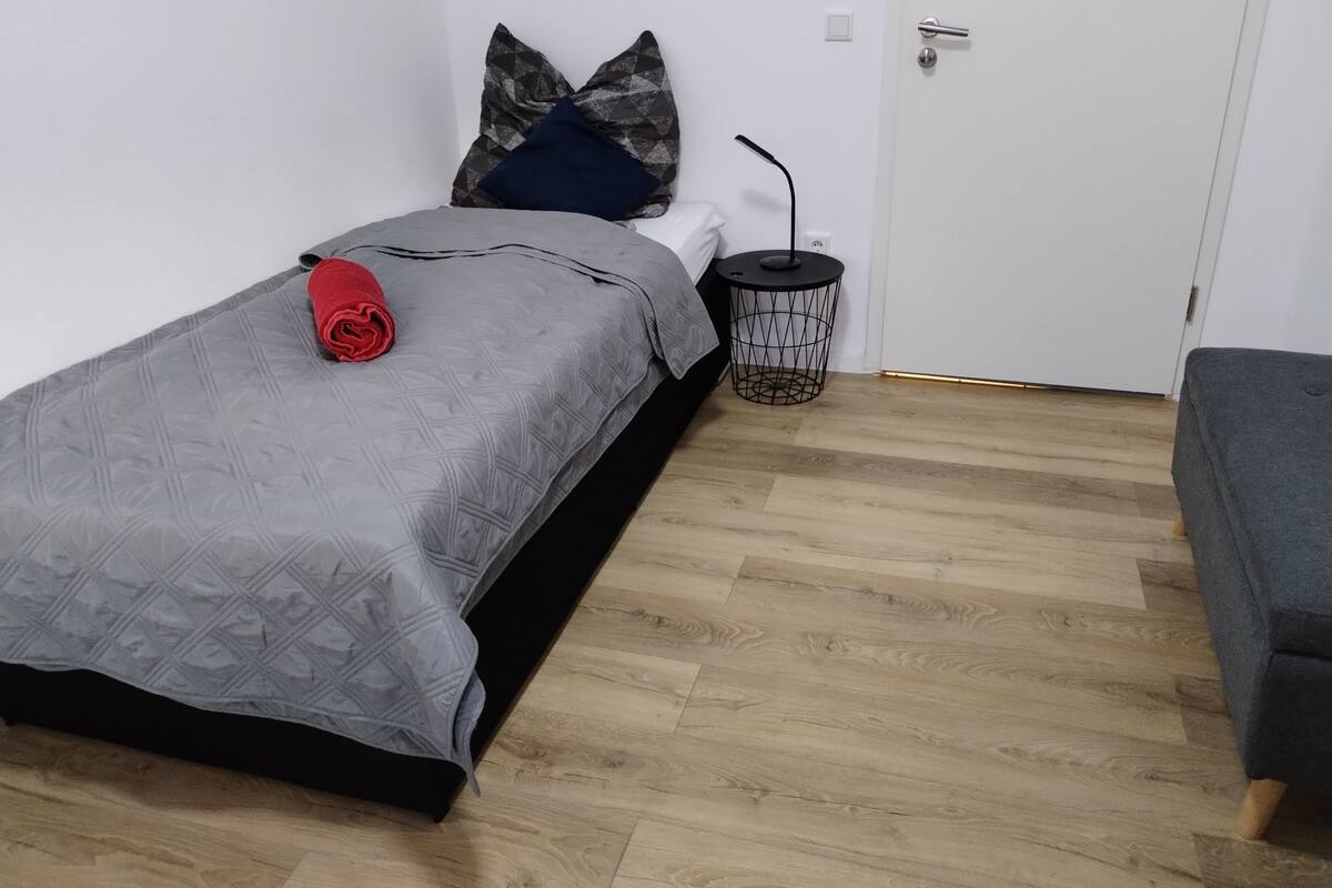 A small, neatly arranged bedroom with a single bed, a bedside table, a waste bin, and a door.