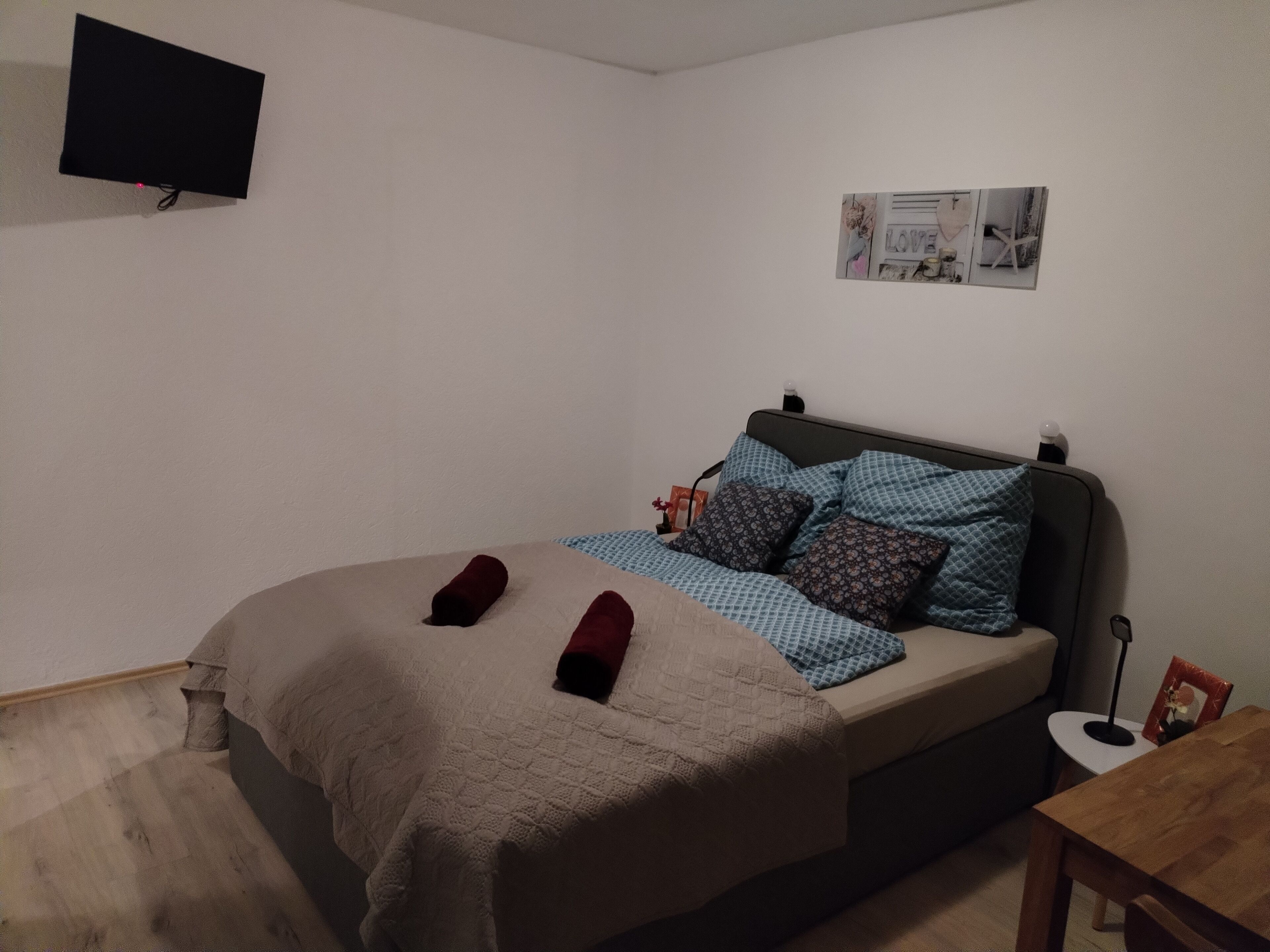 Une chambre à coucher avec un lit, un téléviseur à écran plat fixé au mur et une petite table sur laquelle est posé un cadre photo.