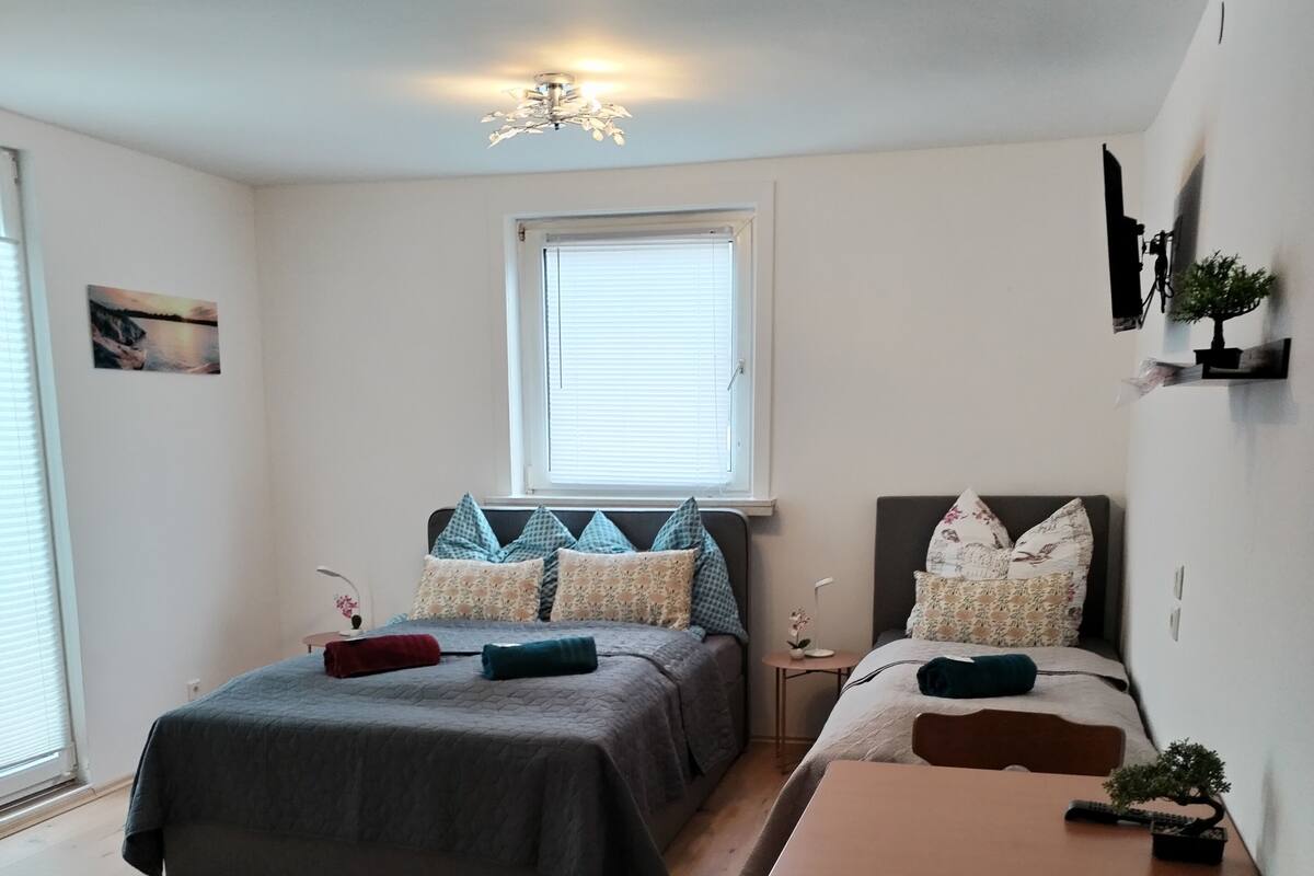 A bedroom with two beds, a wooden table, a chair, a ceiling fan, and a window with blinds.