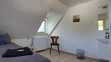A bedroom with a sloped ceiling, a bed, a chair, a radiator, a window, and a small table with a lamp.