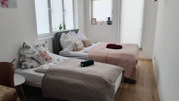 A bedroom with two beds, a small table, and a chair. There are windows with blinds, a wall-mounted shelf with decorative items, and a wooden floor.