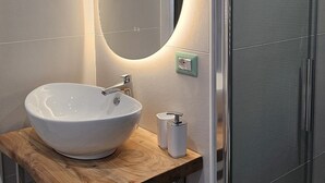 Une salle de bain moderne avec un lavabo blanc, un plan de toilette en bois, un miroir doté d’un éclairage intégré et une cabine de douche en verre.