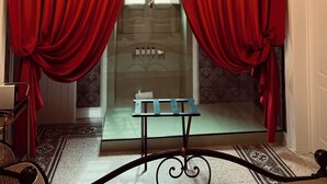 A room with a chandelier, red curtains, a glass door leading to a bathroom, and a patterned floor.