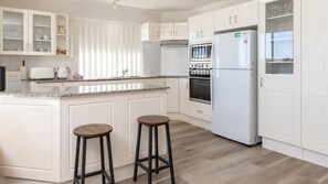 A modern kitchen with white cabinetry, a built-in oven, a refrigerator, and two stools. - Haven Waters 8 on the lakefront (Forster)