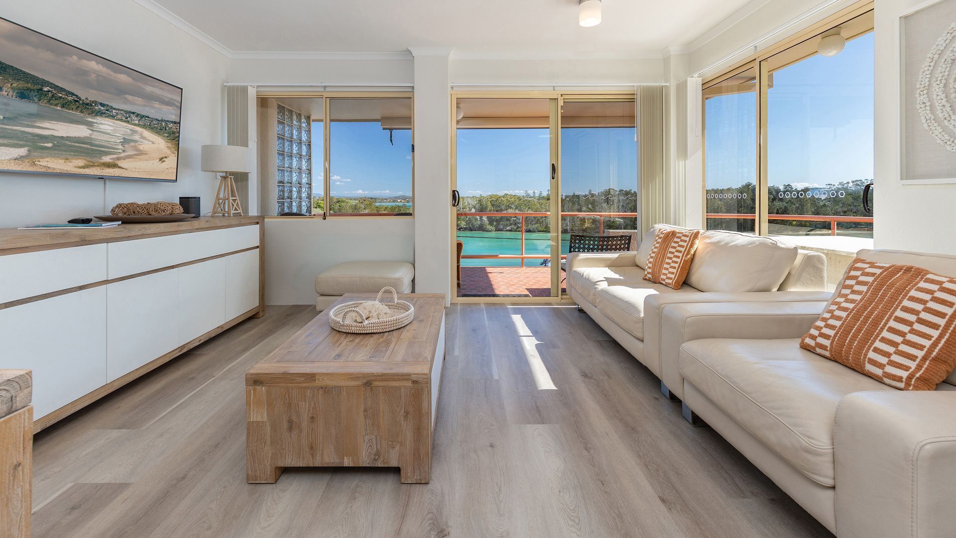 Ein modernes Wohnzimmer mit einem Holztisch, einer Couch mit gemusterten Kissen und einem großen Fenster mit Blick auf Strand und Berge.