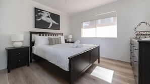 A modern bedroom with a bed, nightstands, a dresser, and a window with blinds. - Haven Waters 8 on the lakefront (Forster)
