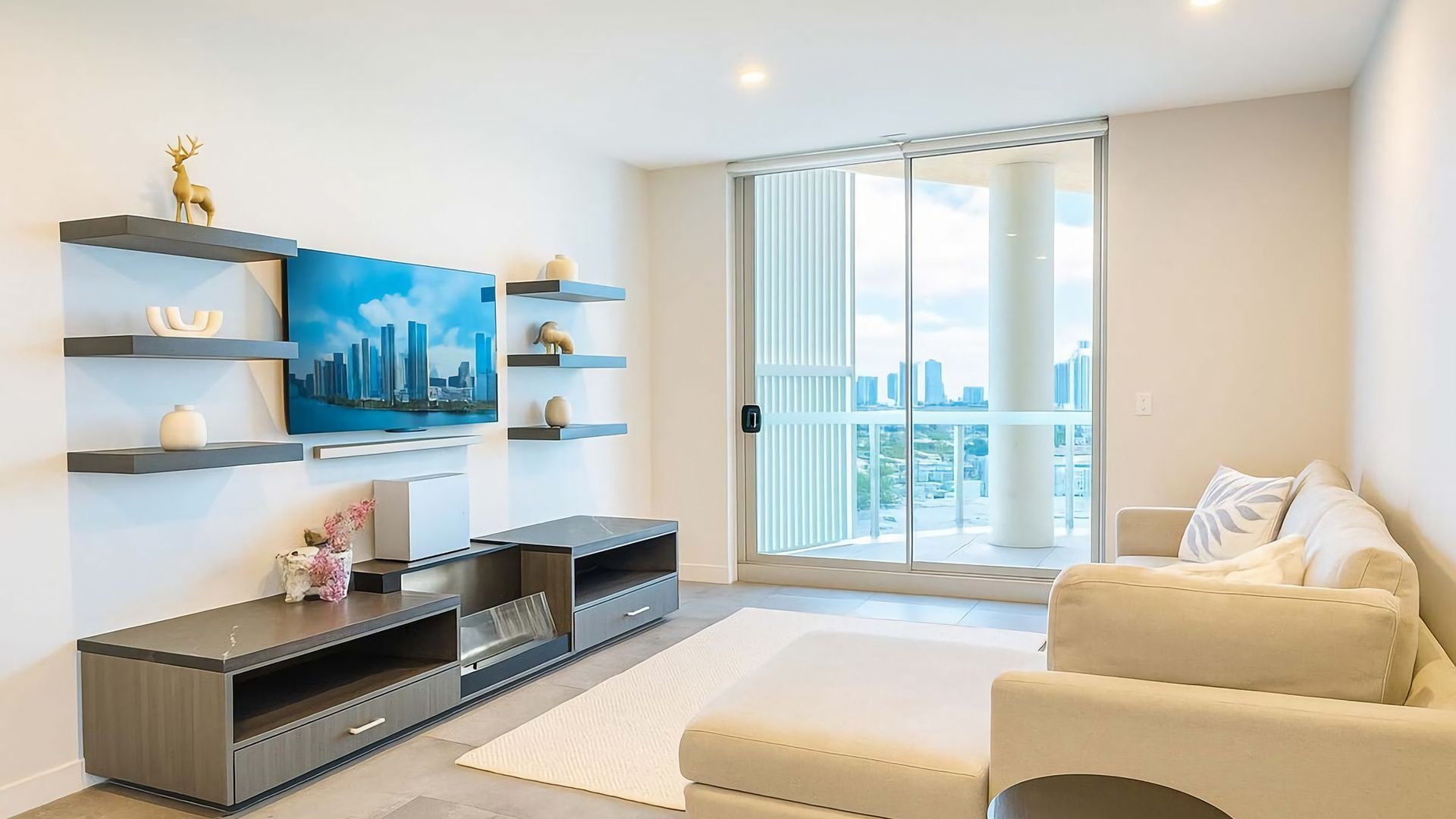 A modern living room with a flat-screen TV, built-in shelves, a sofa, and a large sliding glass door leading to a balcony with a city view.