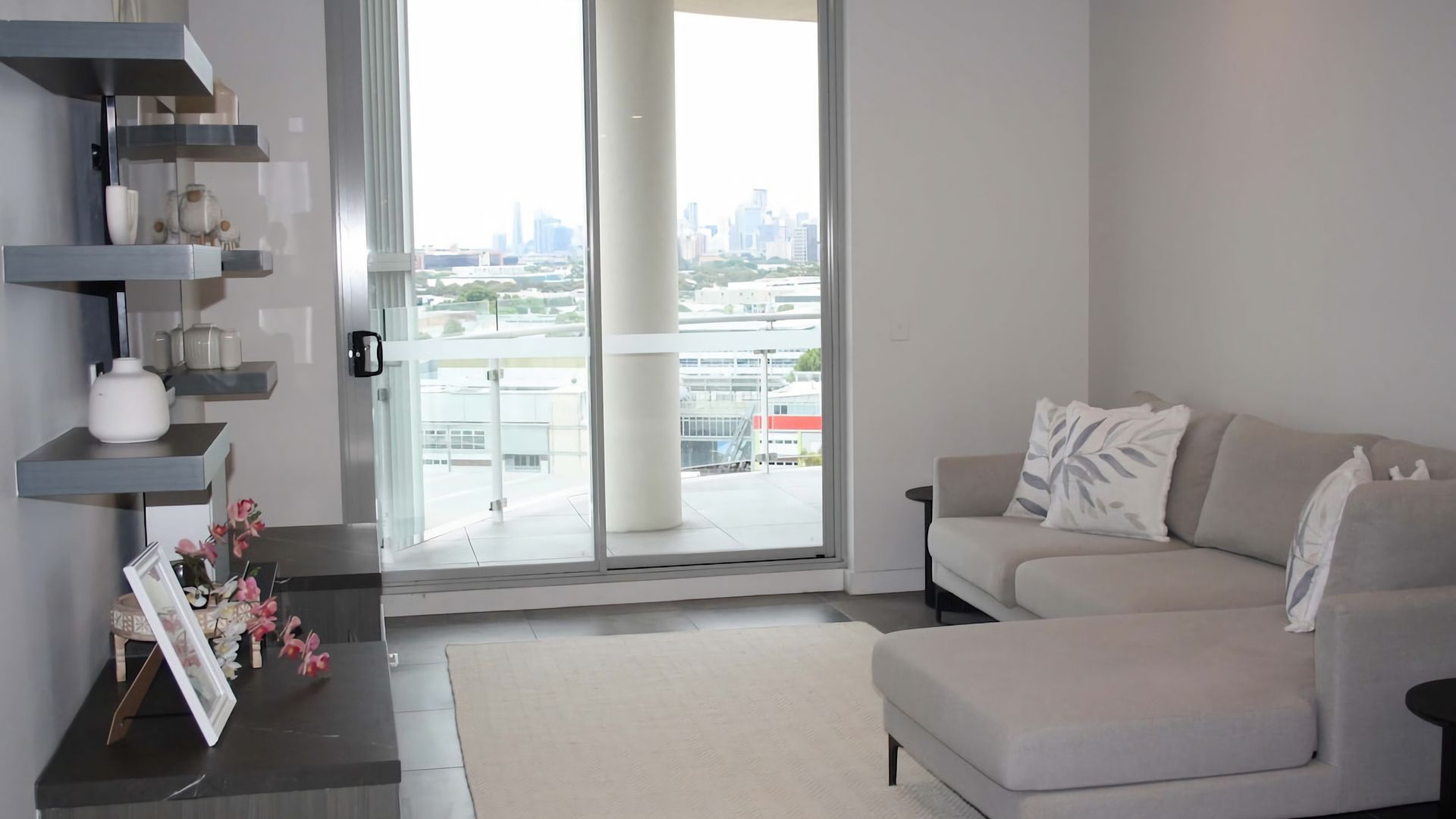 Una sala de estar moderna con un sofá, una mesa de centro y vistas a un paisaje urbano a través de amplios ventanales.