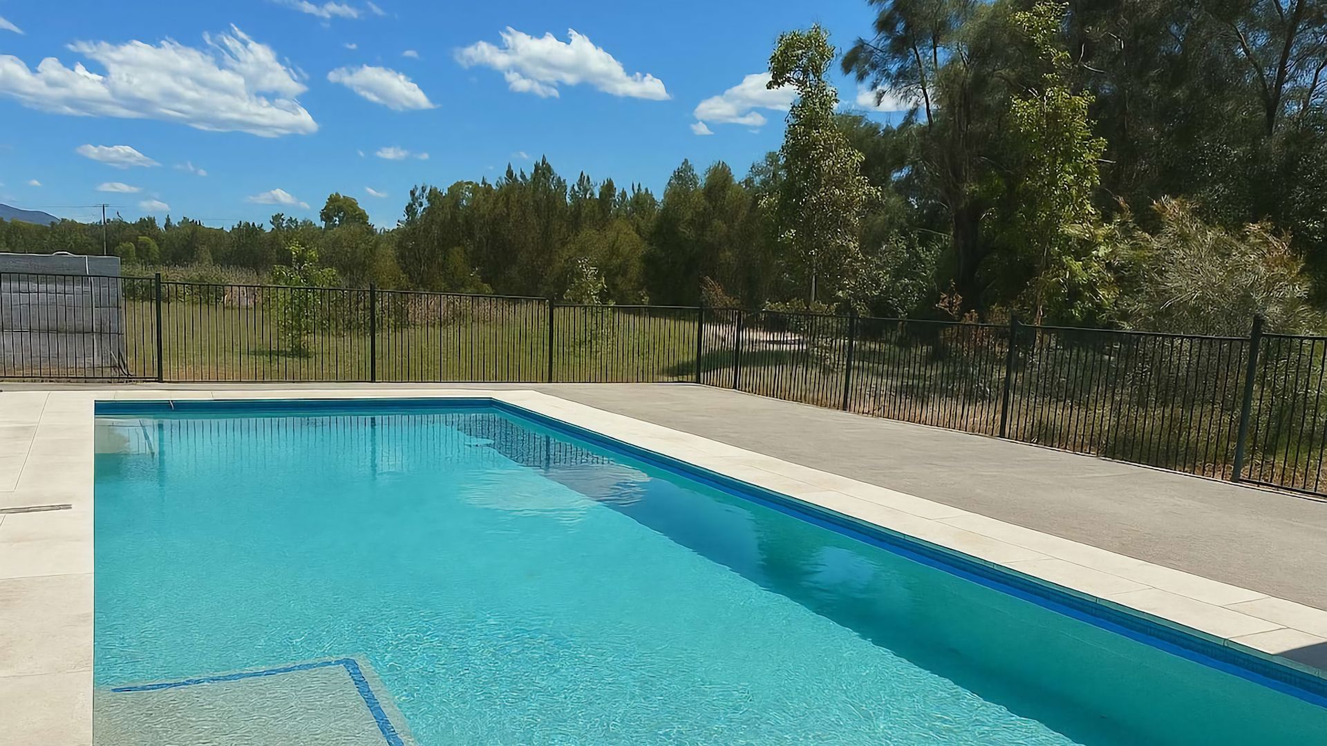 Una piscina rettangolare con acqua cristallina e blu, circondata da un solarium in cemento e una recinzione in metallo nero.