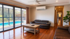 A spacious living room with a large window, a leather sofa, a wooden coffee table, and an air conditioning unit. - Casa Sangiovese | In the Heart of the Hunter Valley (Pokolbin)