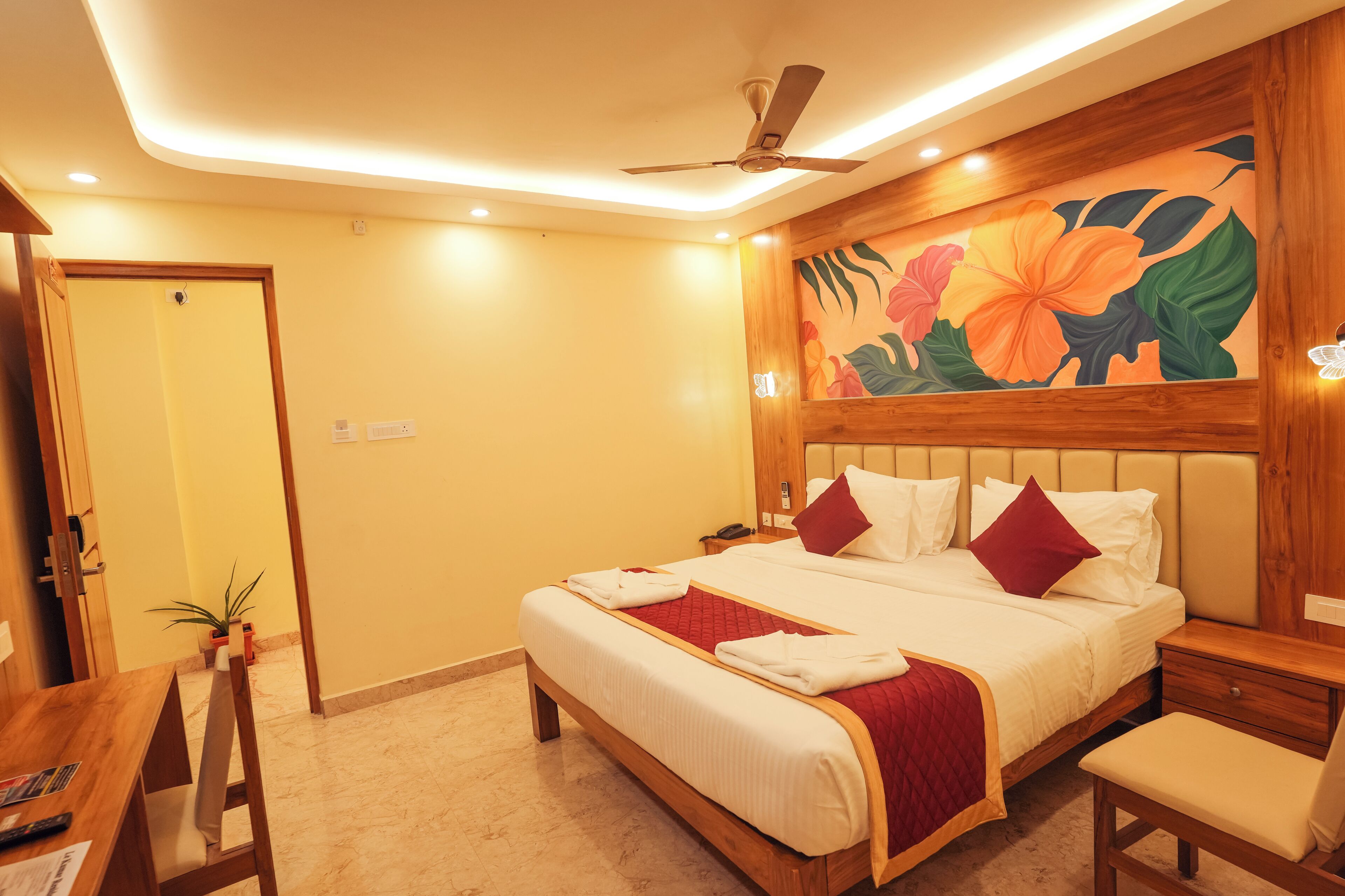 A hotel room with a bed, bedside tables, a desk with a chair, a ceiling fan, and a wall with a floral mural.